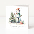 A square Christmas greeting card standing upright on a white surface. The front shows an illustrated snowman wearing a Santa hat and plaid scarf beside a small decorated tree and wrapped gifts, with the text 'Wishing you a Merry Christmas' at the bottom.