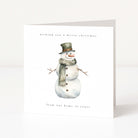 A square white Christmas greeting card standing upright on a white surface. The front features a watercolor-style snowman wearing a dark green top hat and a gray-green scarf, with small black text reading 'wishing you a merry christmas' at the top and 'from our home to yours' at the bottom.