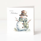 A square white Christmas greeting card standing upright, featuring a watercolor illustration of a snowman wearing a brown hat and a green-and-white striped scarf, with the text 'MERRY Christmas' printed on the left side.