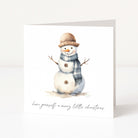A square white Christmas greeting card standing upright on a white surface, printed with a watercolour snowman wearing a tan hat and blue-grey scarf, with the phrase 'have yourself a merry little christmas' in black script at the bottom.