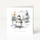 A white Christmas greeting card featuring a watercolour winter illustration of a snowman wearing a black top hat and a red-and-green plaid scarf, standing in a snowy landscape with bare trees, evergreen trees, and a glowing street lamp. The text 'warmest winter wishes' is printed below the illustration.