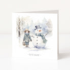 A square Christmas greeting card with a watercolour winter illustration showing a child in a hat and coat standing beside a snowman, with the text '~ let it snow ~' at the bottom, photographed on a clean white background.