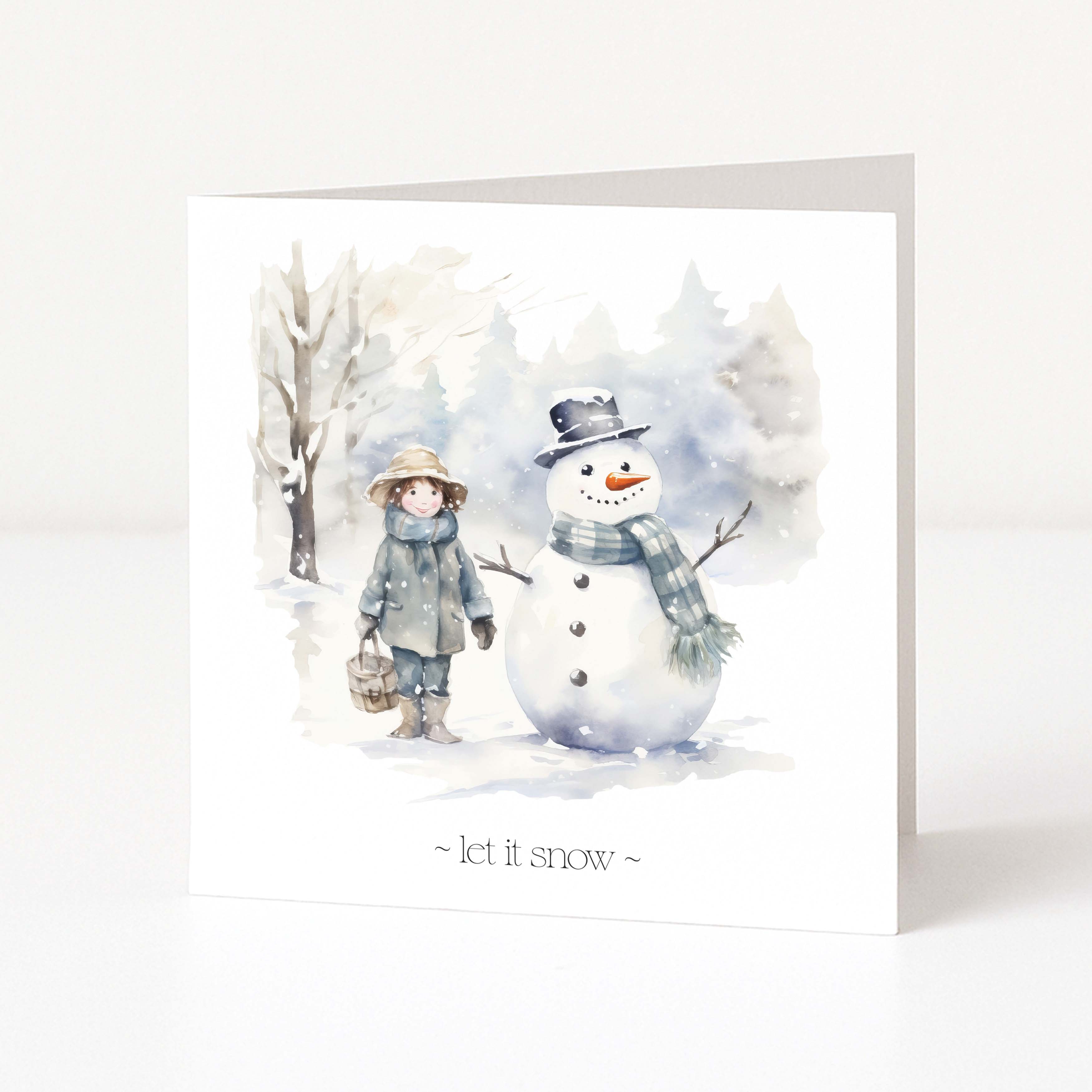 A square Christmas greeting card with a watercolour winter illustration showing a child in a hat and coat standing beside a snowman, with the text '~ let it snow ~' at the bottom, photographed on a clean white background.