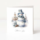 A square white Christmas greeting card standing upright on a white surface, featuring a watercolor illustration of a snowman wearing a top hat and scarf with snowy trees and a small building in the background. The card is printed with the words 'Peace & Love' in black script at the bottom.