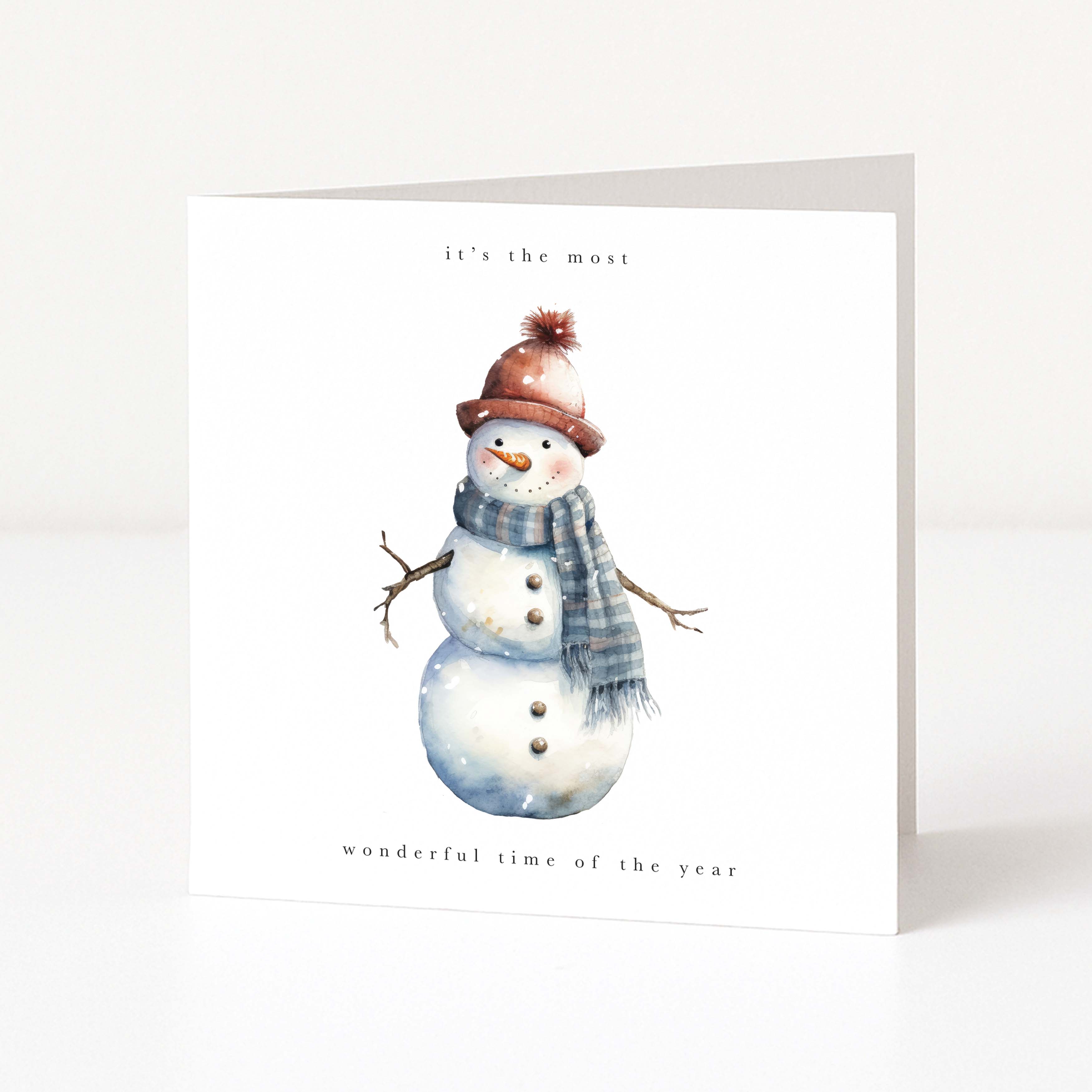 A white Christmas greeting card featuring a watercolour illustration of a snowman wearing a brown knit hat with a pom-pom and a blue-grey striped scarf. The text at the top reads 'it's the most' and the text at the bottom reads 'wonderful time of the year'.