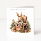 A square greeting card standing upright on a white surface against a light grey background. The front of the card features a watercolour-style illustration of a rabbit sitting in a brown armchair and reading a book, with a small decorated tree, books, and gifts around it.