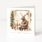 A square, freestanding white holiday card. The watercolour illustration shows an anthropomorphic stag with large antlers sitting comfortably in a wooden chair next to a window. The stag wears a festive red, white, and brown plaid winter sweater and holds a hot drink on a saucer. To the left is a decorated Christmas tree with warm lights and a small side table with a lamp. The text at the bottom reads, "wishing you a WARM and COSY christmas."