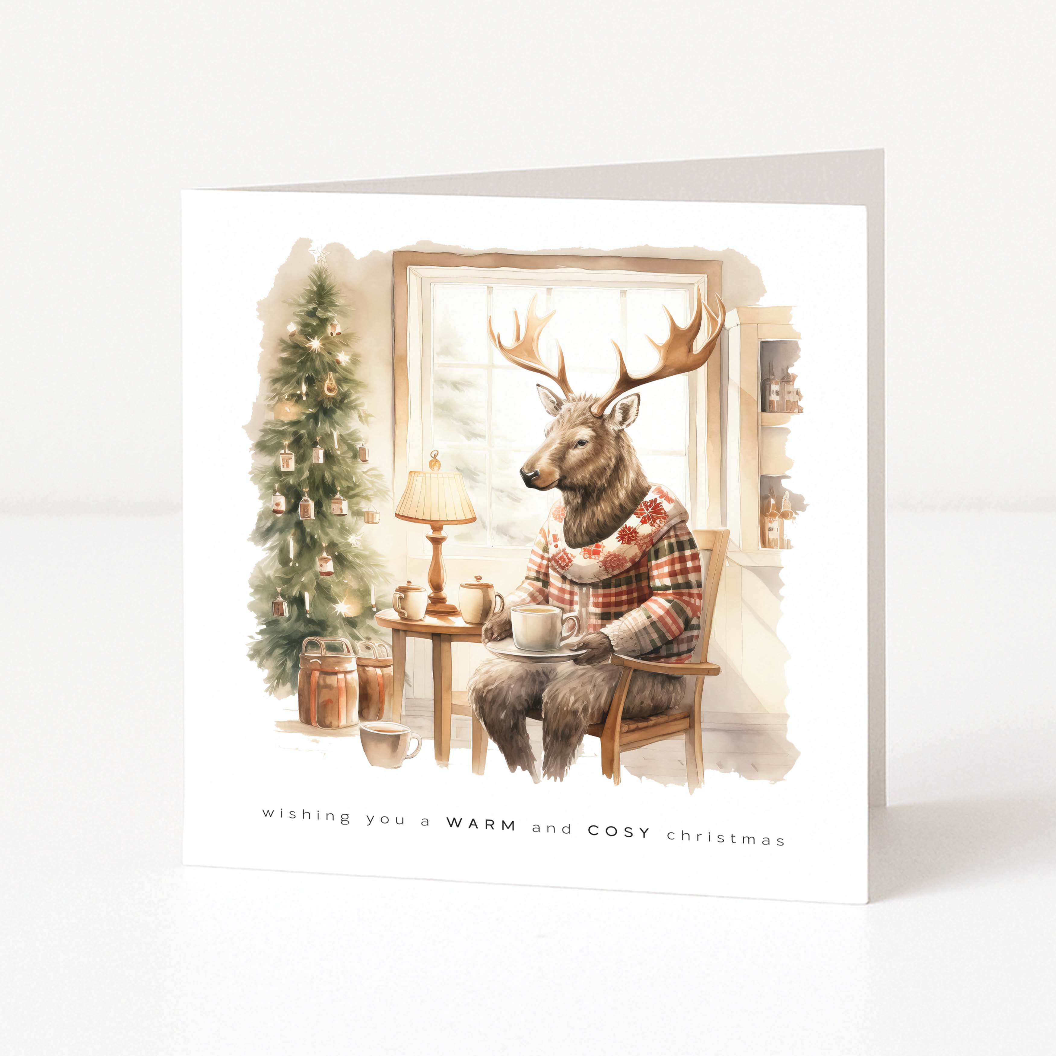A square, freestanding white holiday card. The watercolour illustration shows an anthropomorphic stag with large antlers sitting comfortably in a wooden chair next to a window. The stag wears a festive red, white, and brown plaid winter sweater and holds a hot drink on a saucer. To the left is a decorated Christmas tree with warm lights and a small side table with a lamp. The text at the bottom reads, "wishing you a WARM and COSY christmas."