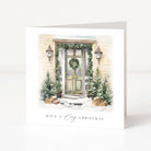 A square white greeting card standing upright on a white surface, printed with an illustrated snowy front door, greenery, lanterns, small trees, and the text 'HAVE A Cozy CHRISTMAS'.