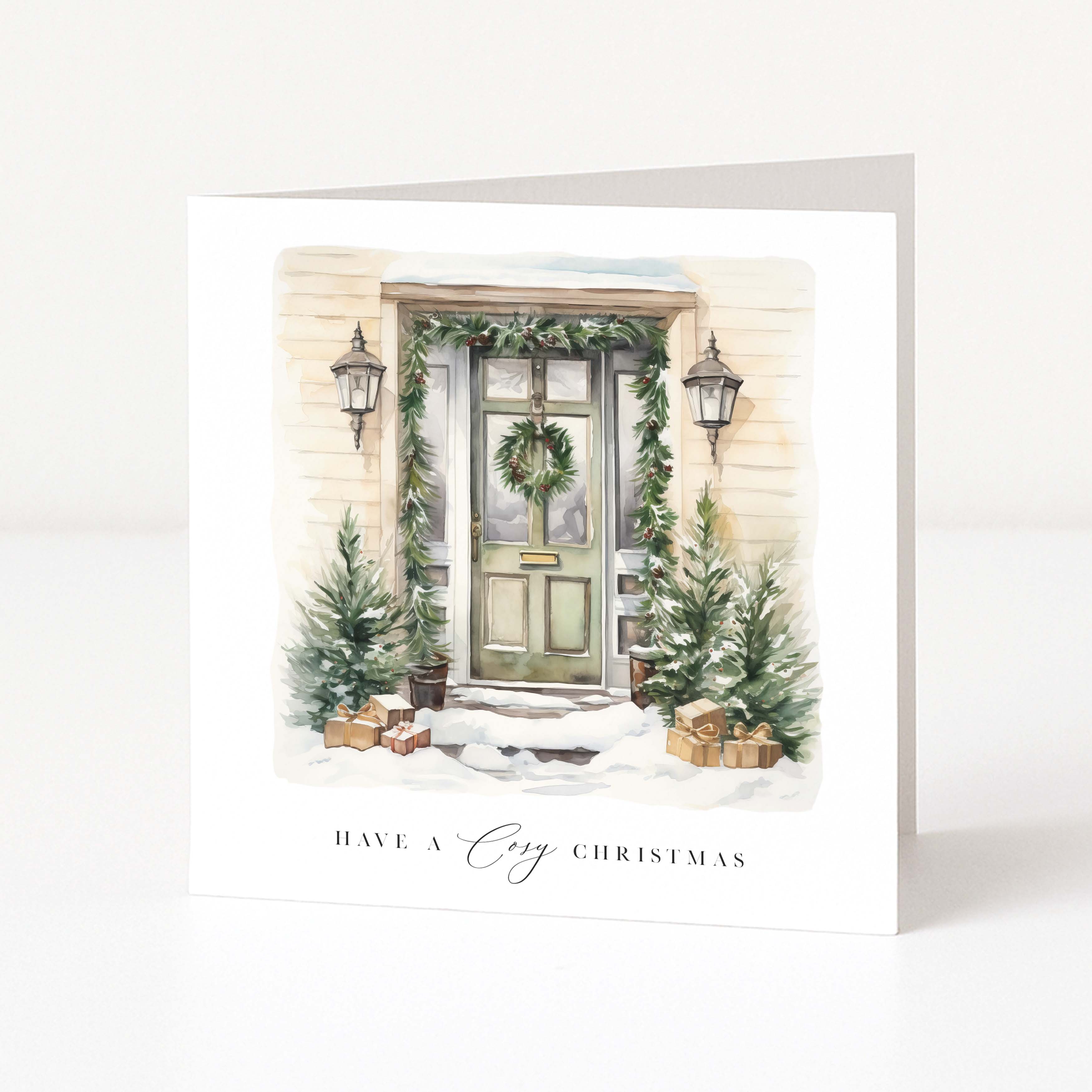 A square white greeting card standing upright on a white surface, printed with an illustrated snowy front door, greenery, lanterns, small trees, and the text 'HAVE A Cozy CHRISTMAS'.