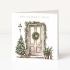 A square greeting card shown standing upright against a clean white background. The front of the card features a watercolor-style illustration of a holiday door scene with a wreath, evergreen garland, small Christmas tree, lamp post, potted plants, and wrapped gifts.
