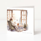 A square Christmas card with a winter illustration showing a person sitting in an armchair by a window, wrapped in a blanket and reading, with a potted tree on the windowsill. The card is photographed against a clean white background.