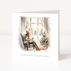 A square greeting card with a white border featuring a watercolor illustration of a woman sitting by a window in a cozy winter scene, with a small Christmas tree and the text 'have a cosy little christmas' at the bottom.
