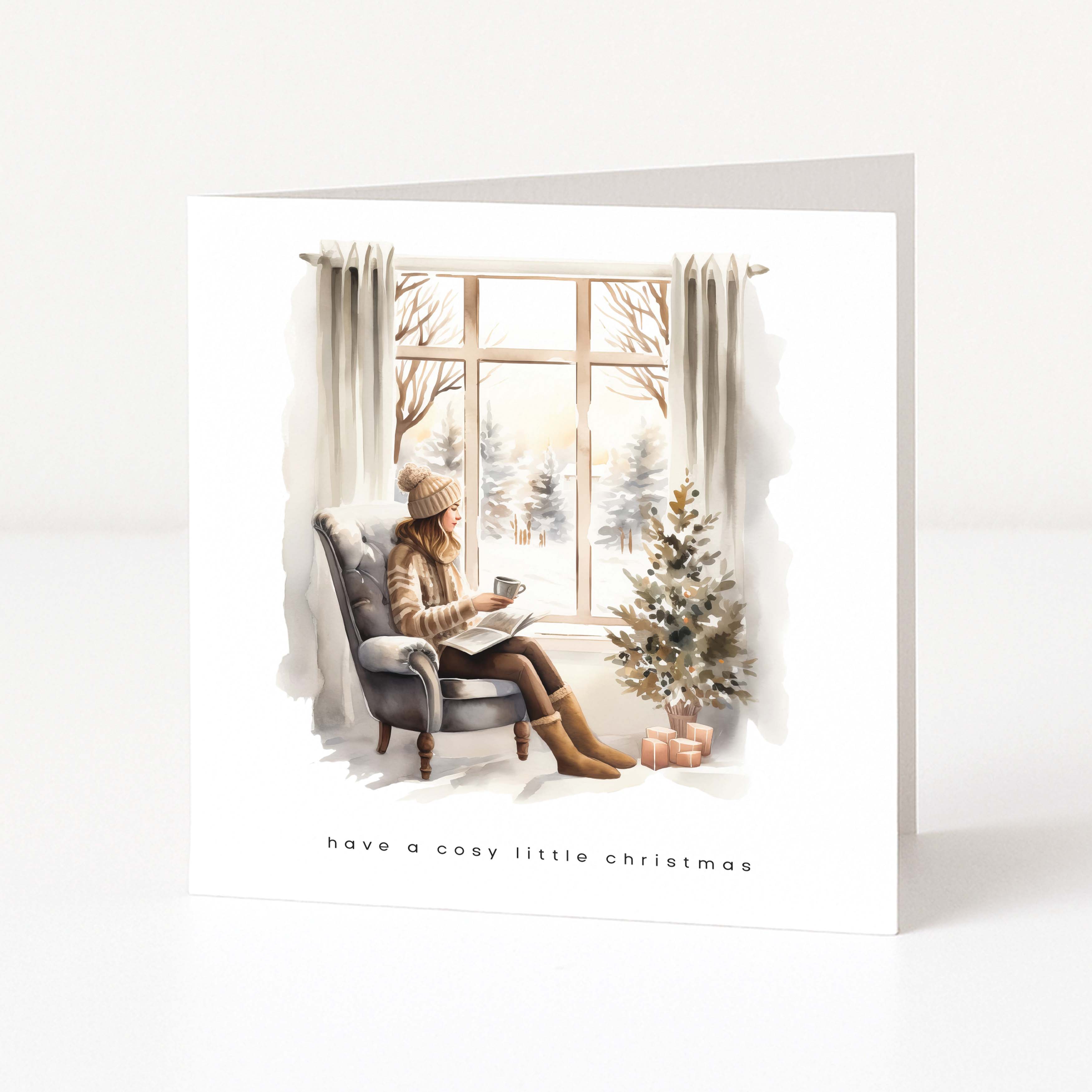 A square greeting card with a white border featuring a watercolor illustration of a woman sitting by a window in a cozy winter scene, with a small Christmas tree and the text 'have a cosy little christmas' at the bottom.