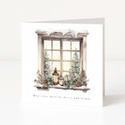 A square Christmas card with a winter window illustration, showing snow on the sill, lanterns, pinecones, evergreen branches, and trees outside the window. The card reads 'May your days be merry and bright!' and is photographed standing upright against a clean white background.
