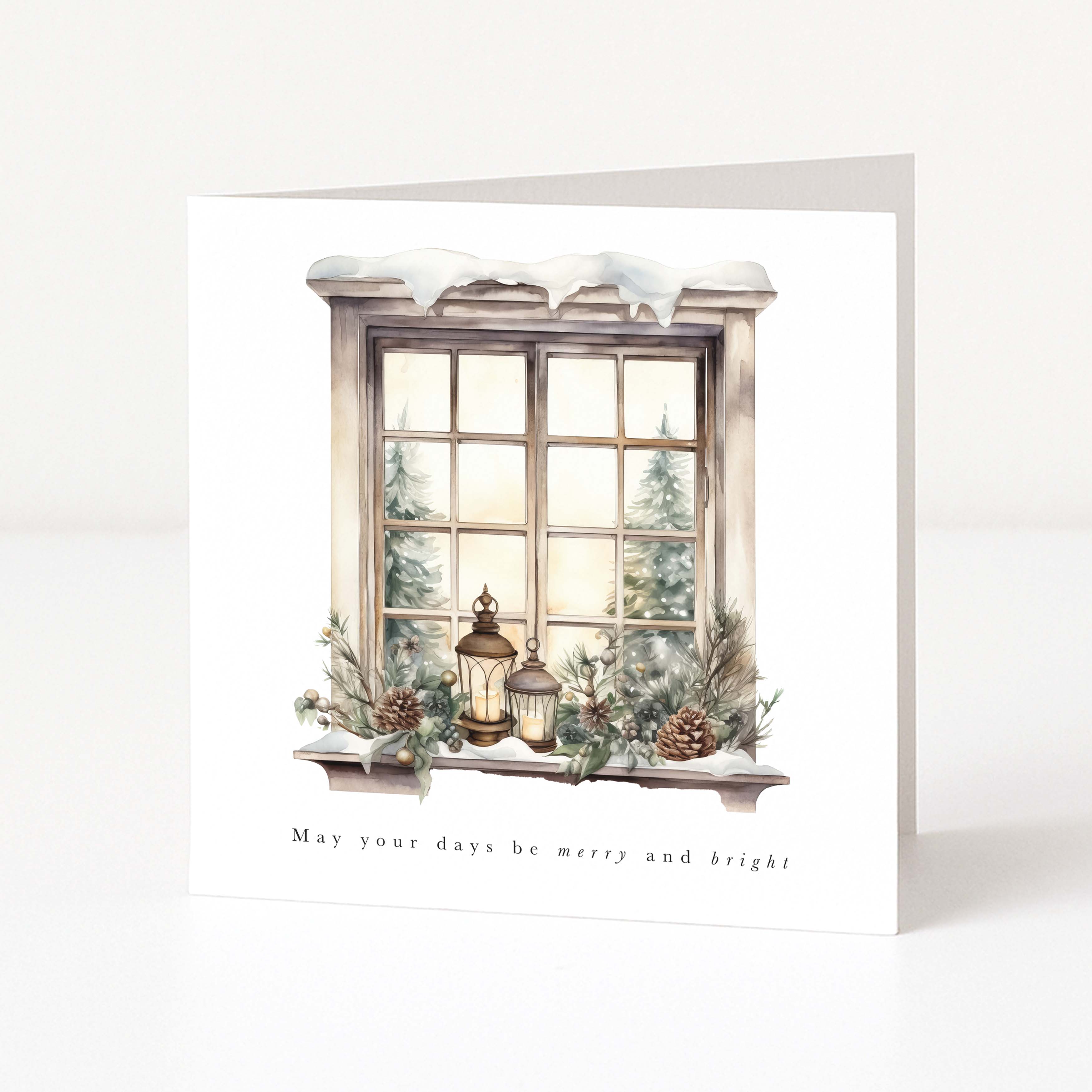 A square Christmas card with a winter window illustration, showing snow on the sill, lanterns, pinecones, evergreen branches, and trees outside the window. The card reads 'May your days be merry and bright!' and is photographed standing upright against a clean white background.