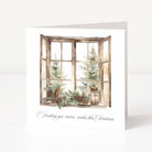 A square white Christmas greeting card standing upright on a white surface, featuring an illustrated open window with Christmas trees, pinecones, greenery, and a lantern, plus the text 'Sending you warm wishes this Christmas' in black script.