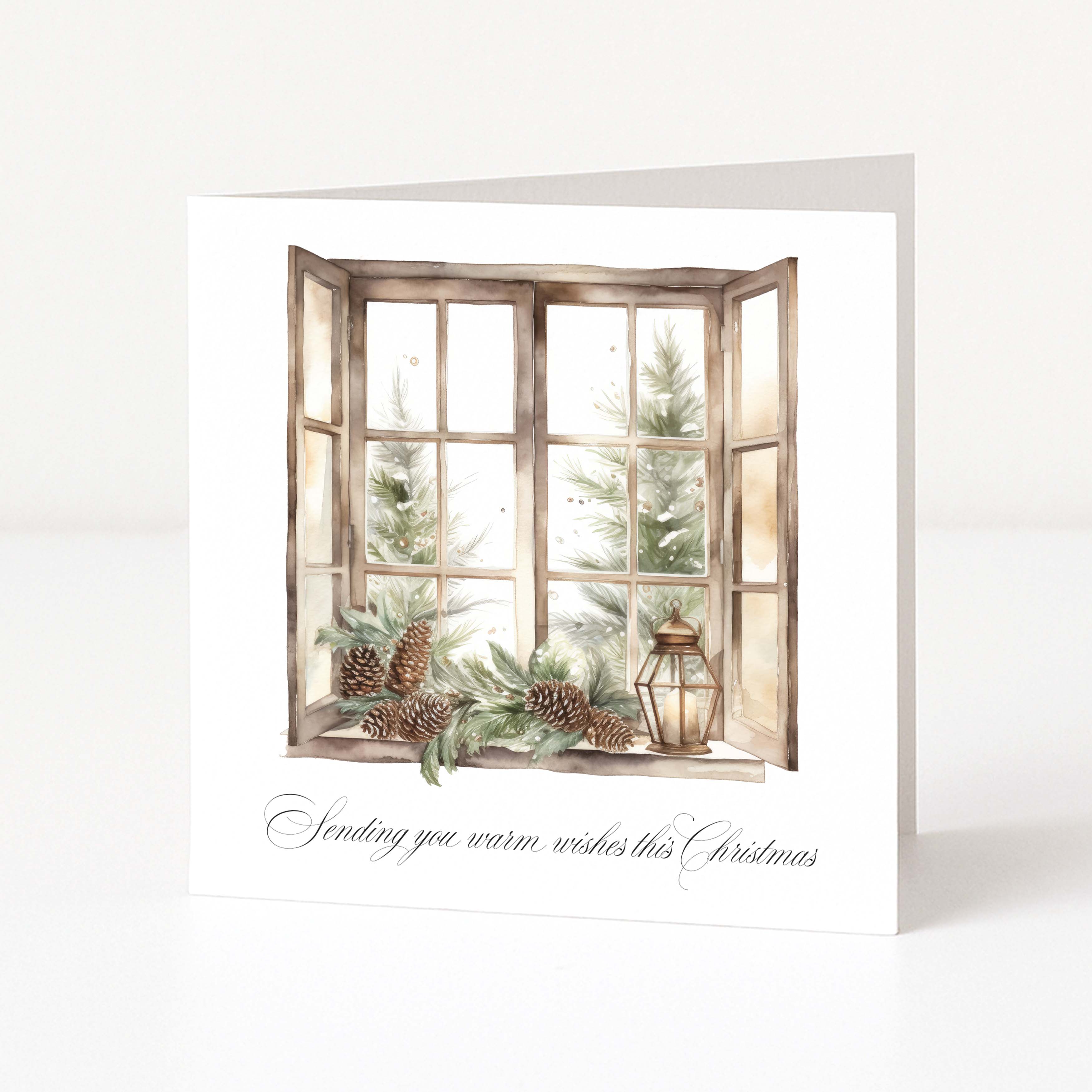 A square white Christmas greeting card standing upright on a white surface, featuring an illustrated open window with Christmas trees, pinecones, greenery, and a lantern, plus the text 'Sending you warm wishes this Christmas' in black script.