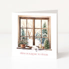 A white Christmas greeting card standing upright, printed with an illustration of a snowy window scene and the red text 'FROM OUR HOME TO YOURS' at the bottom, photographed on a clean white background.