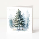 A square Christmas greeting card with a watercolour illustration of a snow-covered evergreen tree in a misty winter forest. Small printed text appears near the upper left, and the card is shown standing upright against a clean white background.