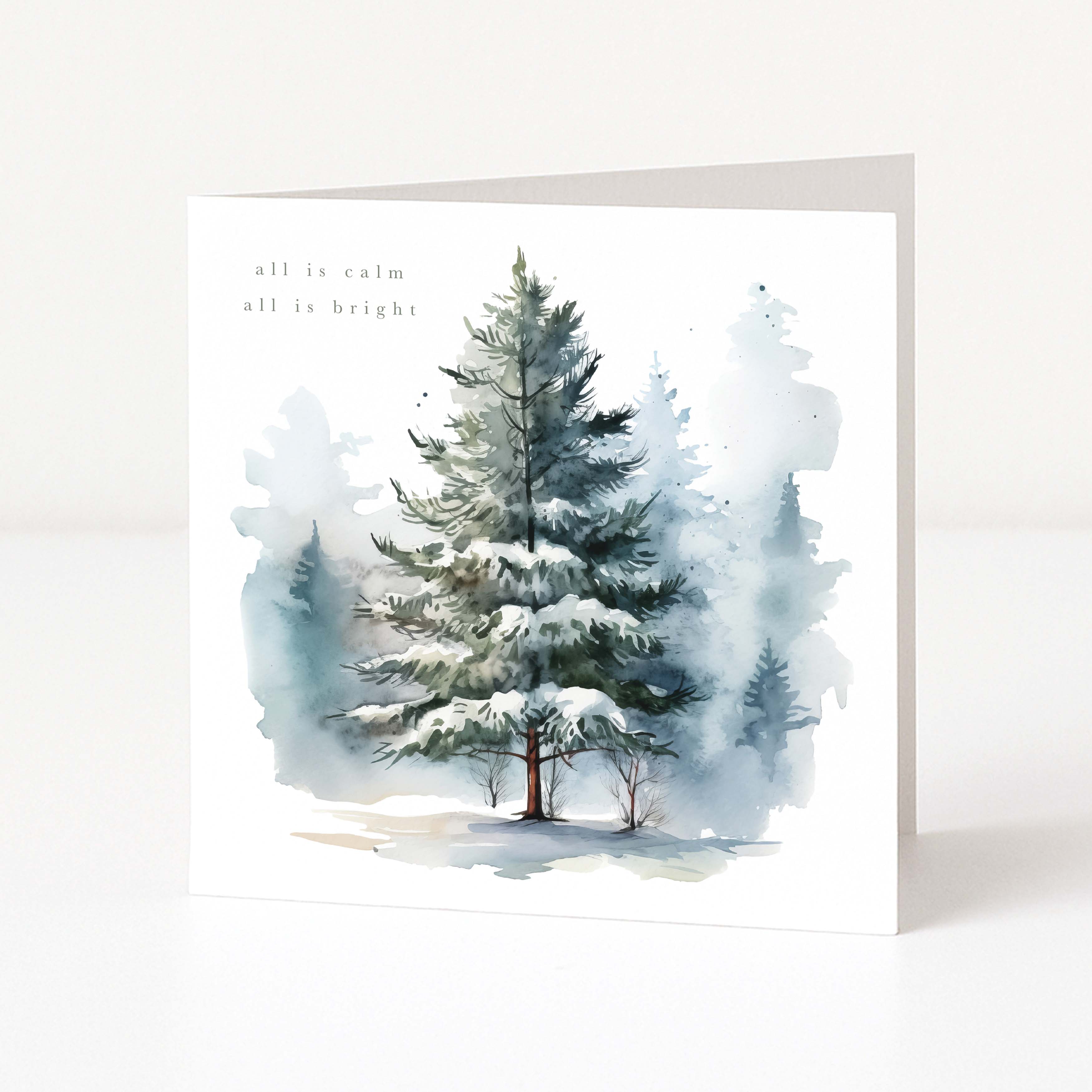 A square Christmas greeting card with a watercolour illustration of a snow-covered evergreen tree in a misty winter forest. Small printed text appears near the upper left, and the card is shown standing upright against a clean white background.