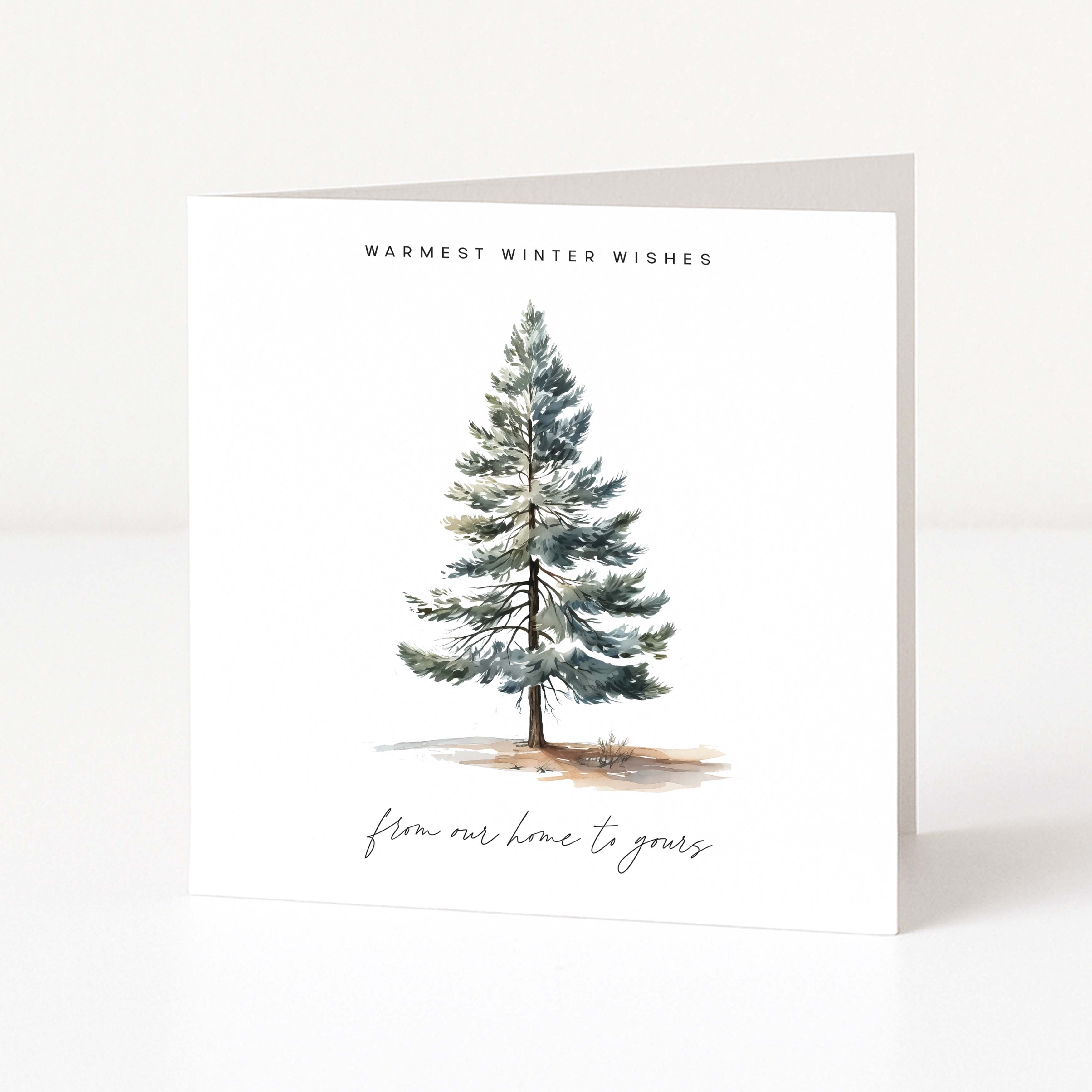 A white Christmas greeting card standing upright on a light surface, featuring a watercolour-style evergreen tree with the text 'WARMEST WINTER WISHES' above and 'from our home to yours' below.