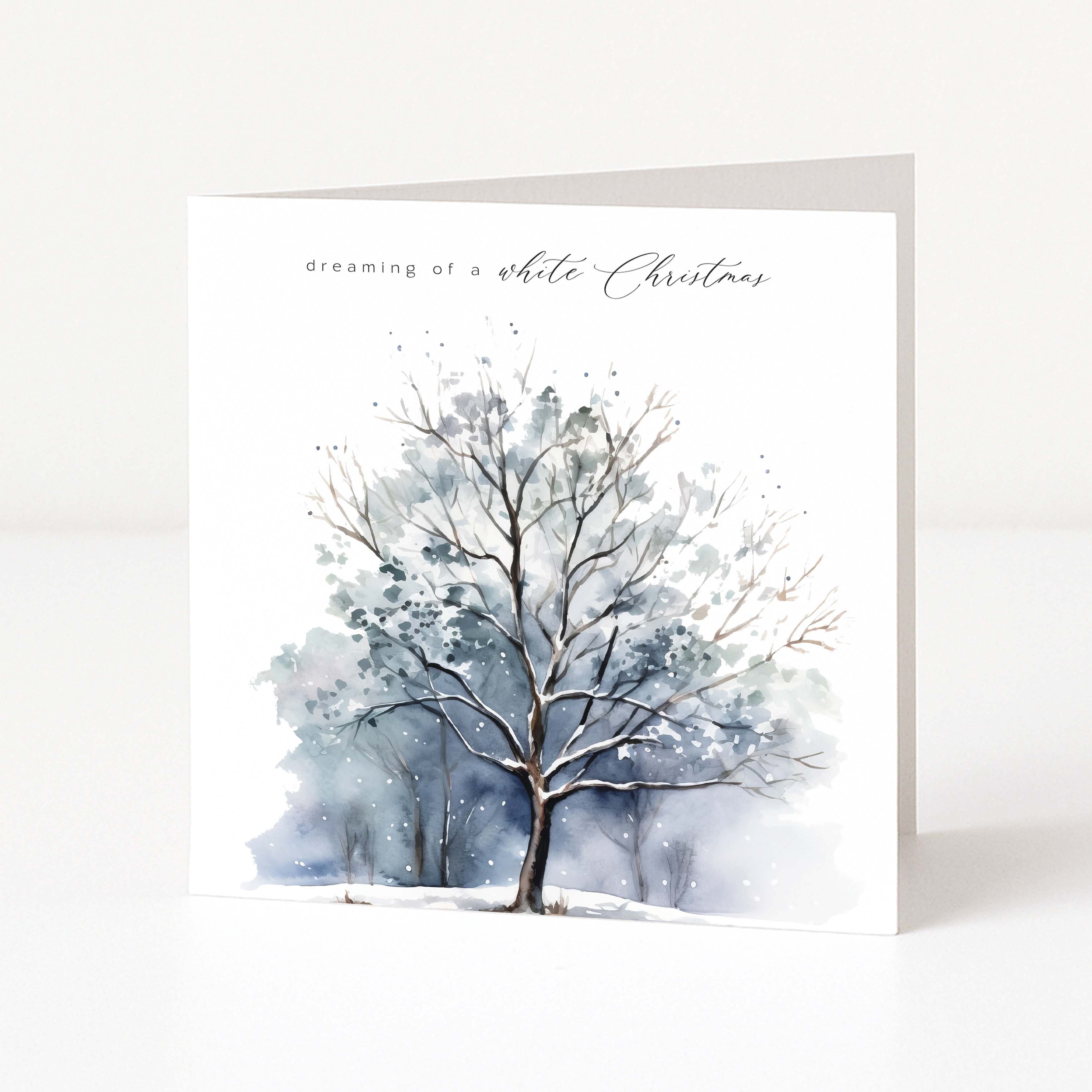 A square Christmas greeting card standing upright on a white surface, featuring a watercolour winter tree illustration with the text 'dreaming of a white Christmas' near the top.