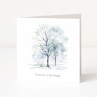 A white Christmas greeting card standing upright, featuring a watercolour illustration of two bare trees in soft blue and grey tones, with the text 'Season's Greetings' printed below.