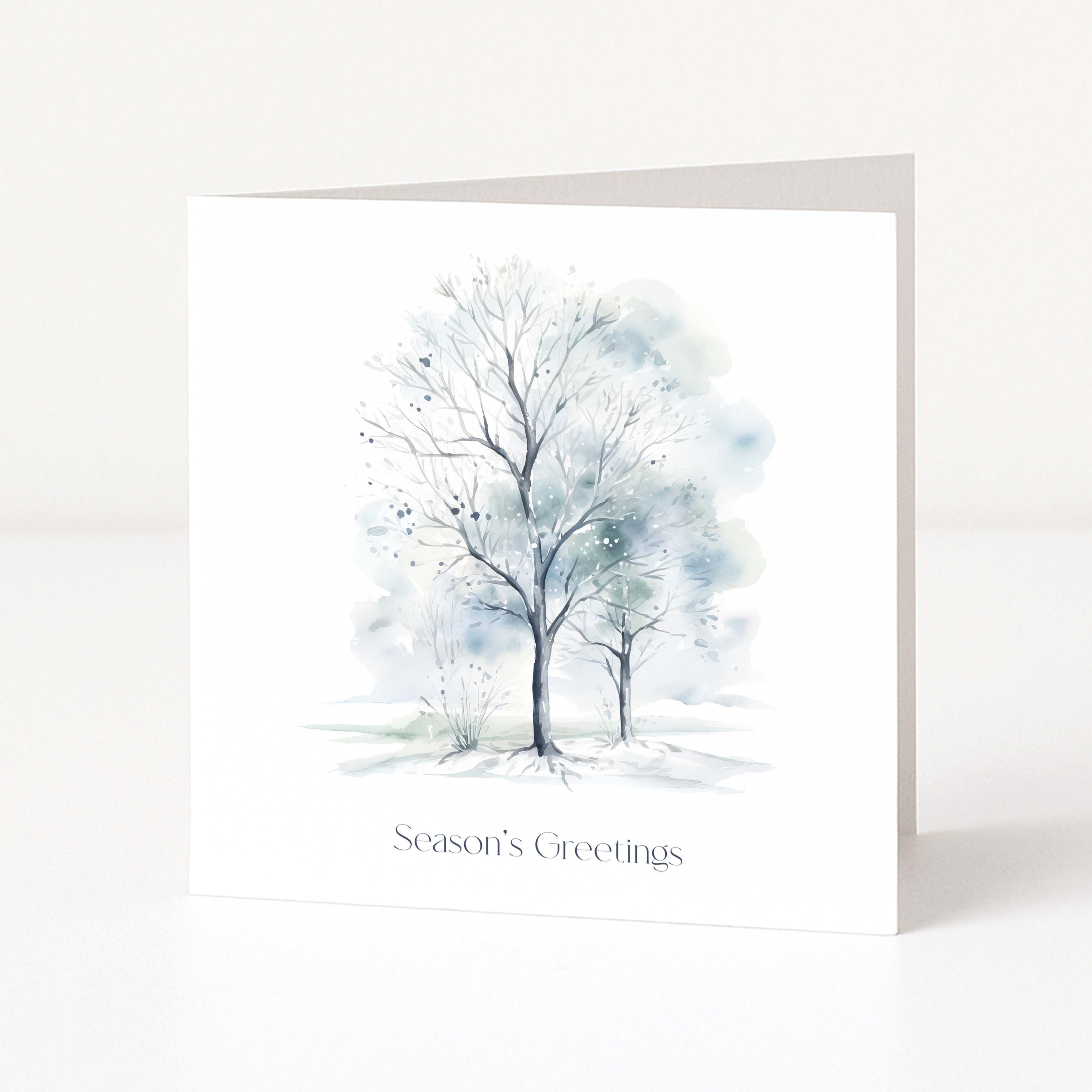A white Christmas greeting card standing upright, featuring a watercolour illustration of two bare trees in soft blue and grey tones, with the text 'Season's Greetings' printed below.