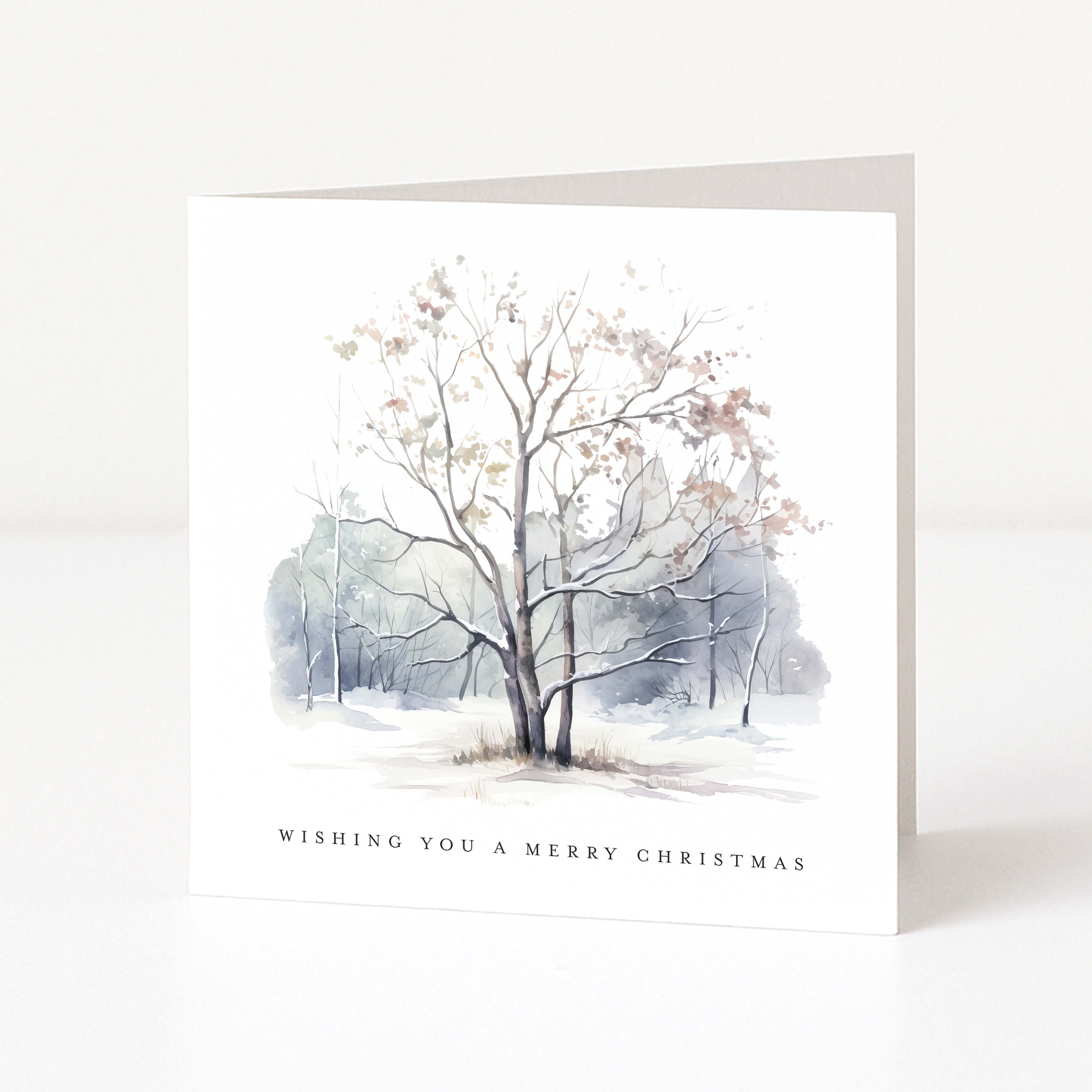 A square white Christmas greeting card standing upright on a white surface, featuring a watercolour winter tree scene in soft grey, blue, and beige tones, with the text 'WISHING YOU A MERRY CHRISTMAS' printed near the bottom.