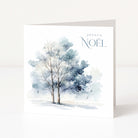 A white Christmas greeting card standing upright, featuring a watercolour painting of two snow-covered trees in soft blue and grey tones, with the text 'JOYEUX NOÉL' printed in the upper right.
