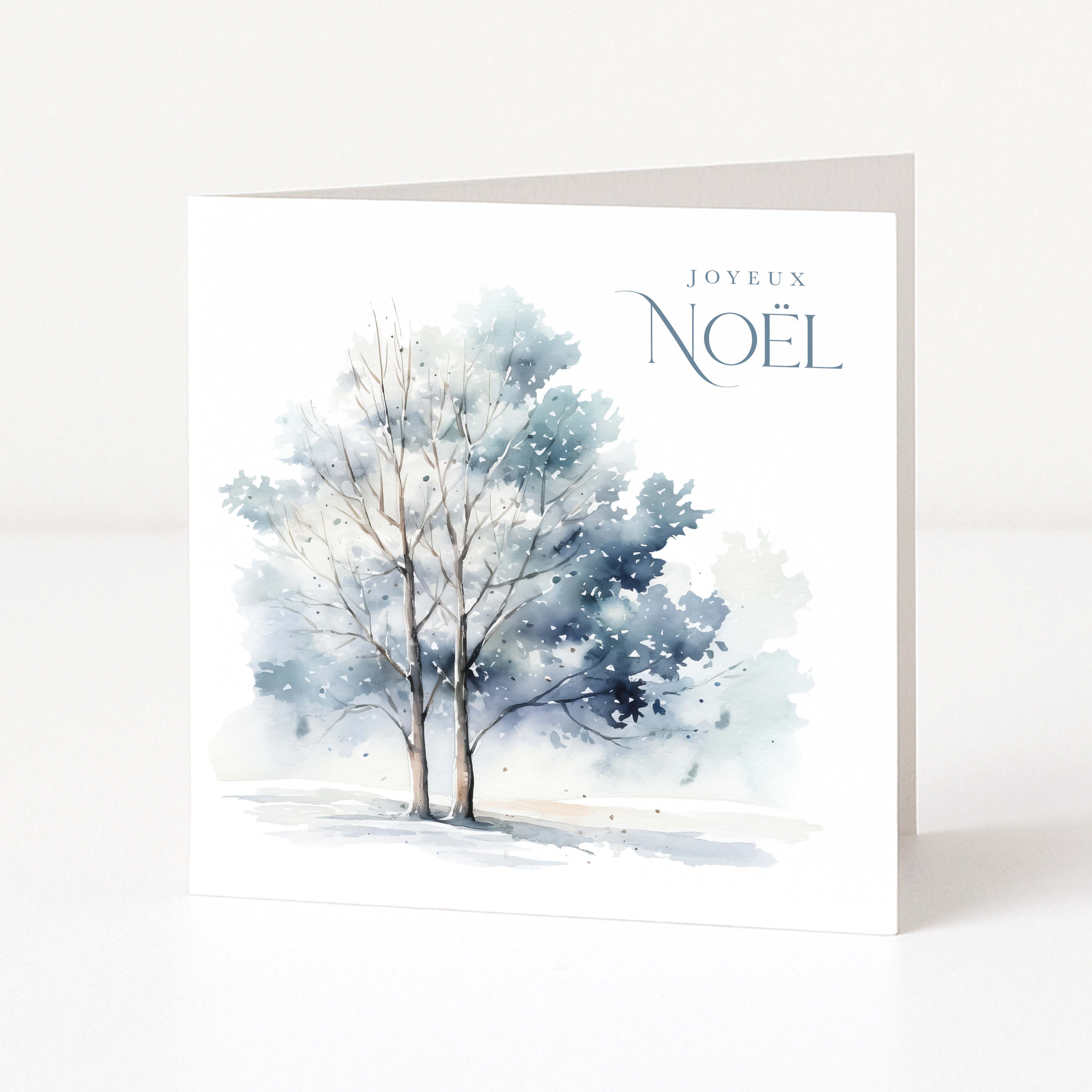 A white Christmas greeting card standing upright, featuring a watercolour painting of two snow-covered trees in soft blue and grey tones, with the text 'JOYEUX NOÉL' printed in the upper right.