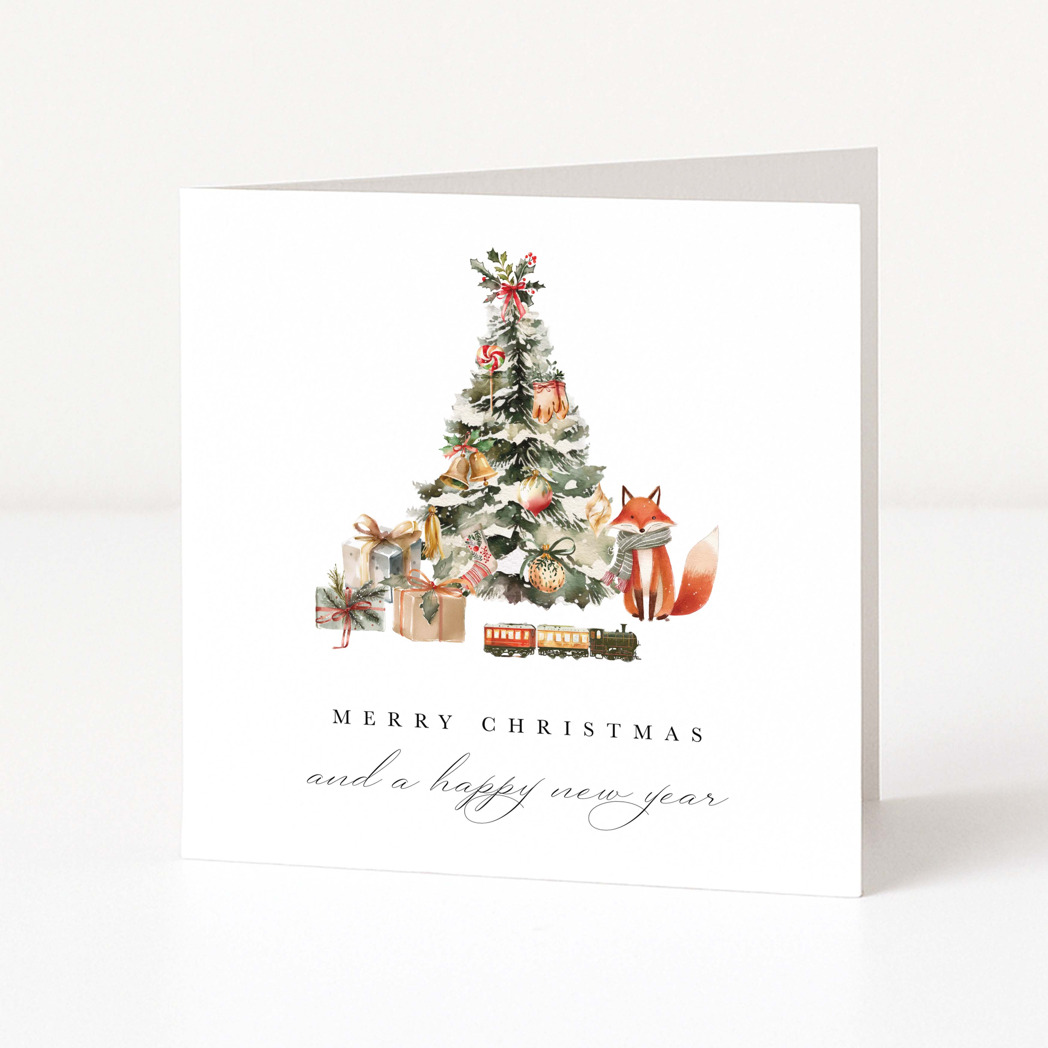 A white square Christmas greeting card standing upright on a white surface, featuring a watercolour-style Christmas tree with ornaments, wrapped gifts, a small train, and a fox. The text reads 'MERRY CHRISTMAS and a happy new year'.