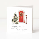 A square white Christmas greeting card standing upright on a light gray surface. The front features an illustration of a red door with a green wreath, a decorated Christmas tree, and a vintage street lamp, with the text 'MERRY CHRISTMAS and a happy new year' below.