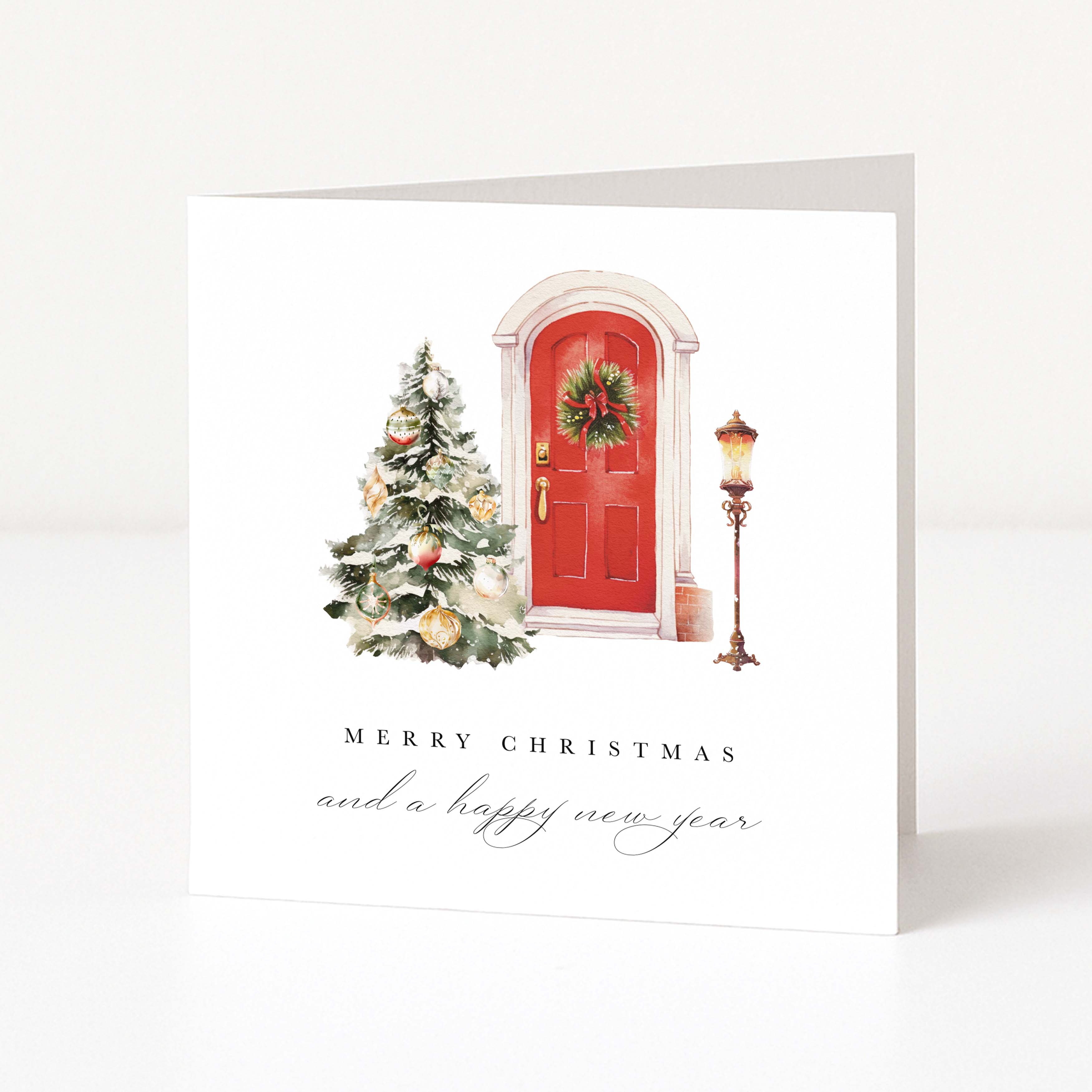 A square white Christmas greeting card standing upright on a light gray surface. The front features an illustration of a red door with a green wreath, a decorated Christmas tree, and a vintage street lamp, with the text 'MERRY CHRISTMAS and a happy new year' below.