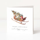 A white Christmas greeting card standing upright, featuring a watercolor illustration of a sleigh carrying a decorated Christmas tree and wrapped gifts, with the text 'MERRY CHRISTMAS and a happy new year' below.