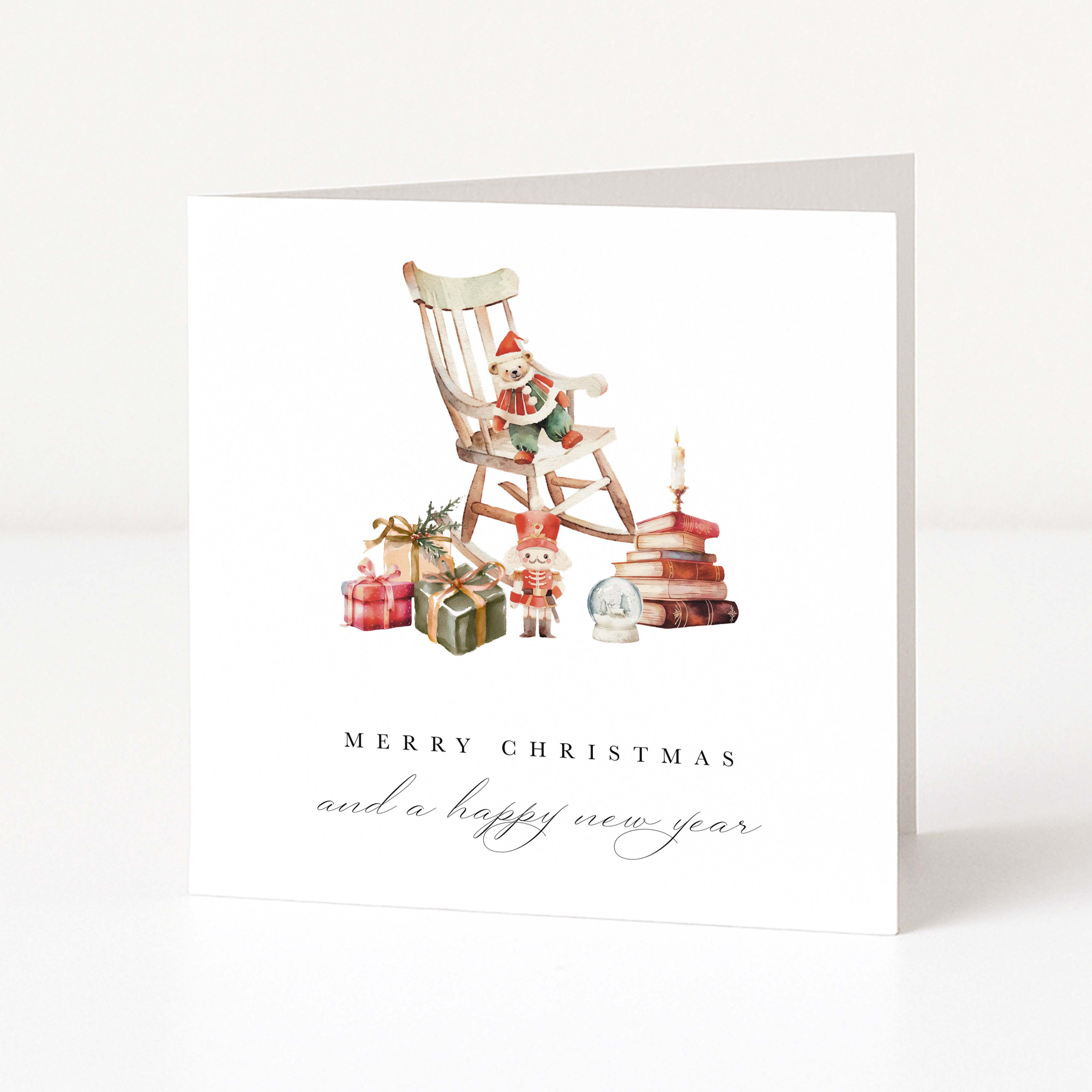 A white greeting card standing upright on a light surface, featuring a watercolour illustration of a Christmas scene with a rocking chair, gifts, books, and a nutcracker. The text reads 'MERRY CHRISTMAS and a happy new year'.