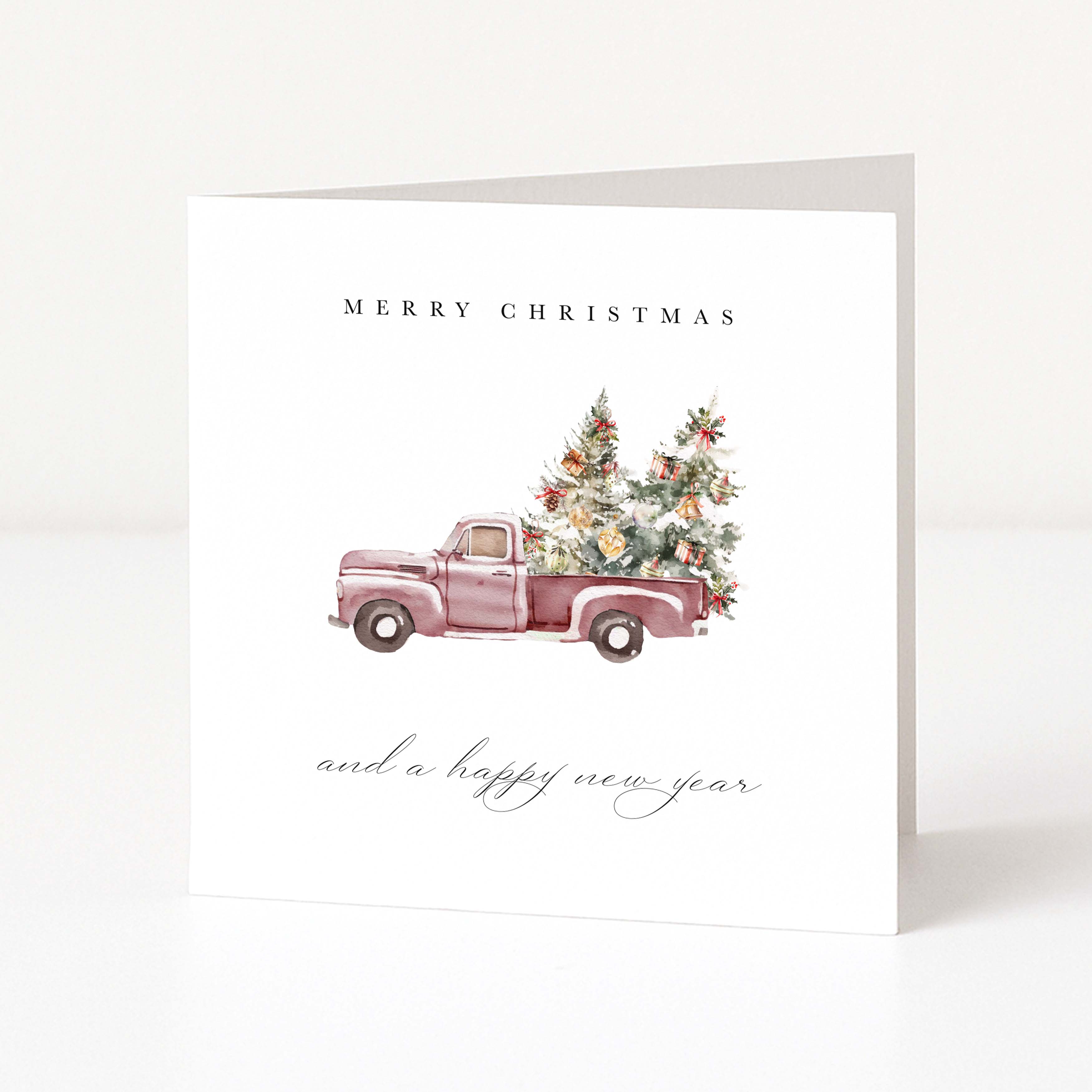 A white Christmas greeting card featuring a watercolour illustration of a vintage pink pickup truck carrying two decorated Christmas trees. The text 'MERRY CHRISTMAS' appears above the truck, and 'and a happy new year' is written below in a cursive font.