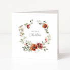 A square white Christmas greeting card with a watercolour floral wreath in muted red, orange, and green tones, plus a small bird and the words 'Merry Christmas' in the centre, shown standing against a clean white background.