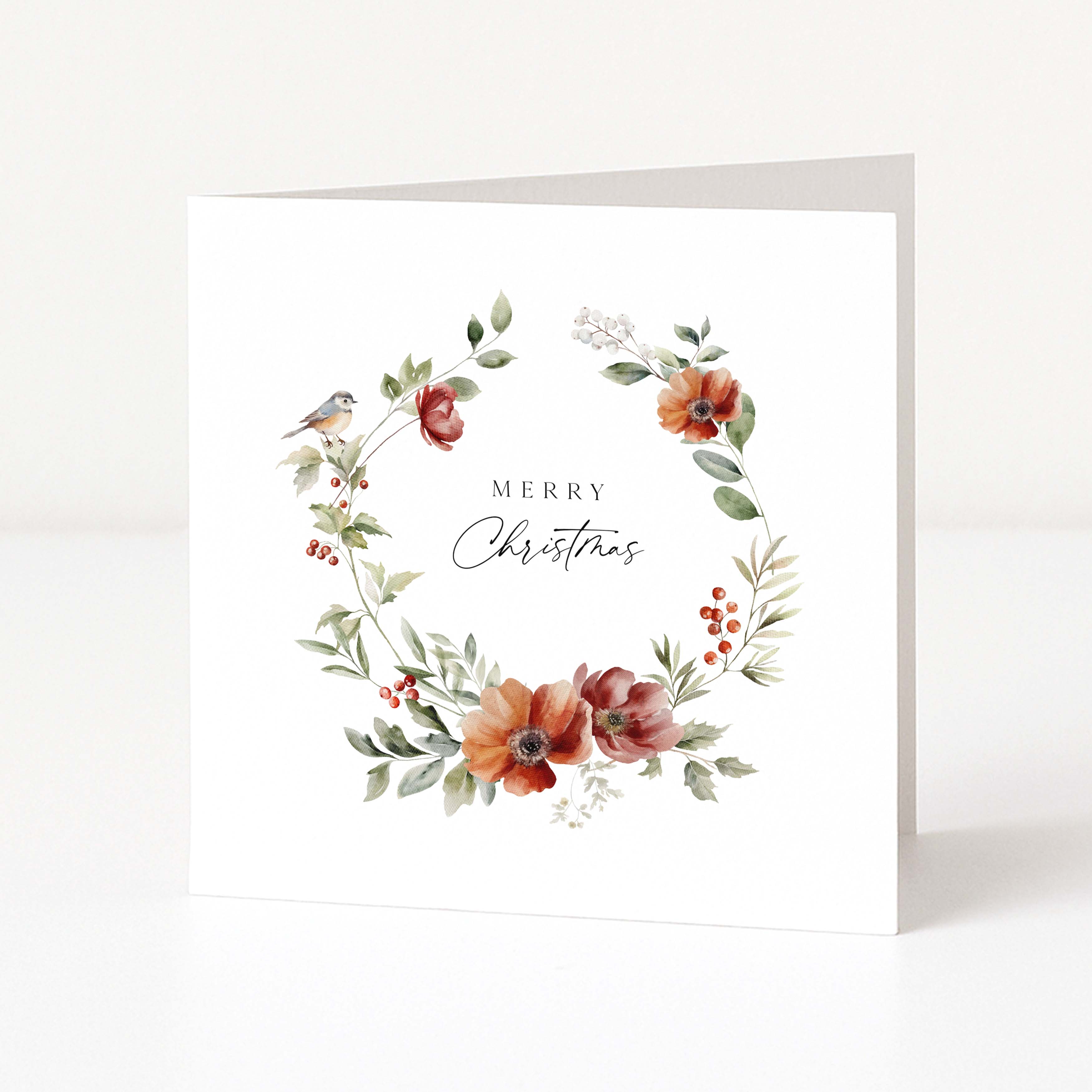 A square white Christmas greeting card with a watercolour floral wreath in muted red, orange, and green tones, plus a small bird and the words 'Merry Christmas' in the centre, shown standing against a clean white background.