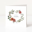A white square Christmas greeting card standing upright on a white surface, featuring a watercolour floral wreath with green leaves, red and orange flowers, and the words “Merry Christmas” in the centre.