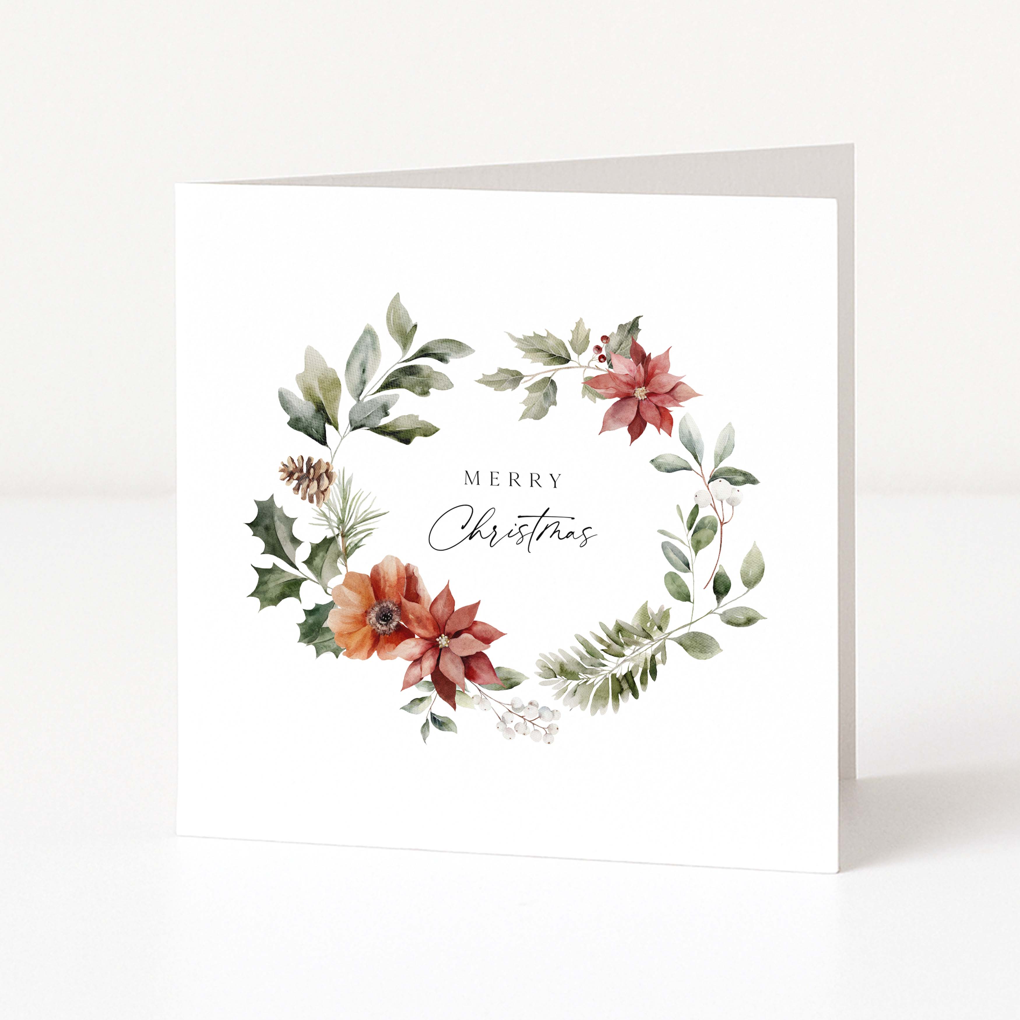 A white square Christmas greeting card standing upright on a white surface, featuring a watercolour floral wreath with green leaves, red and orange flowers, and the words “Merry Christmas” in the centre.
