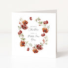 A square white greeting card standing upright on a white surface, featuring a watercolour floral wreath in muted red, rust, and olive tones around the centred text 'Merry Christmas and a Happy New Year.'.