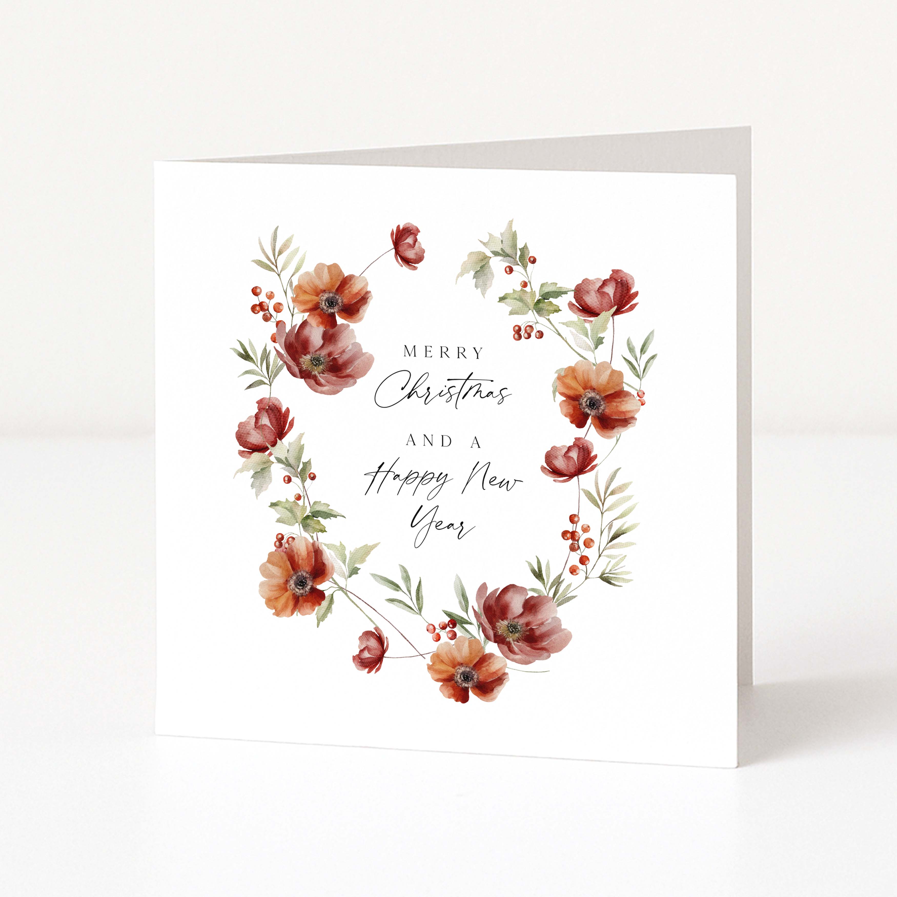 A square white greeting card standing upright on a white surface, featuring a watercolour floral wreath in muted red, rust, and olive tones around the centred text 'Merry Christmas and a Happy New Year.'.
