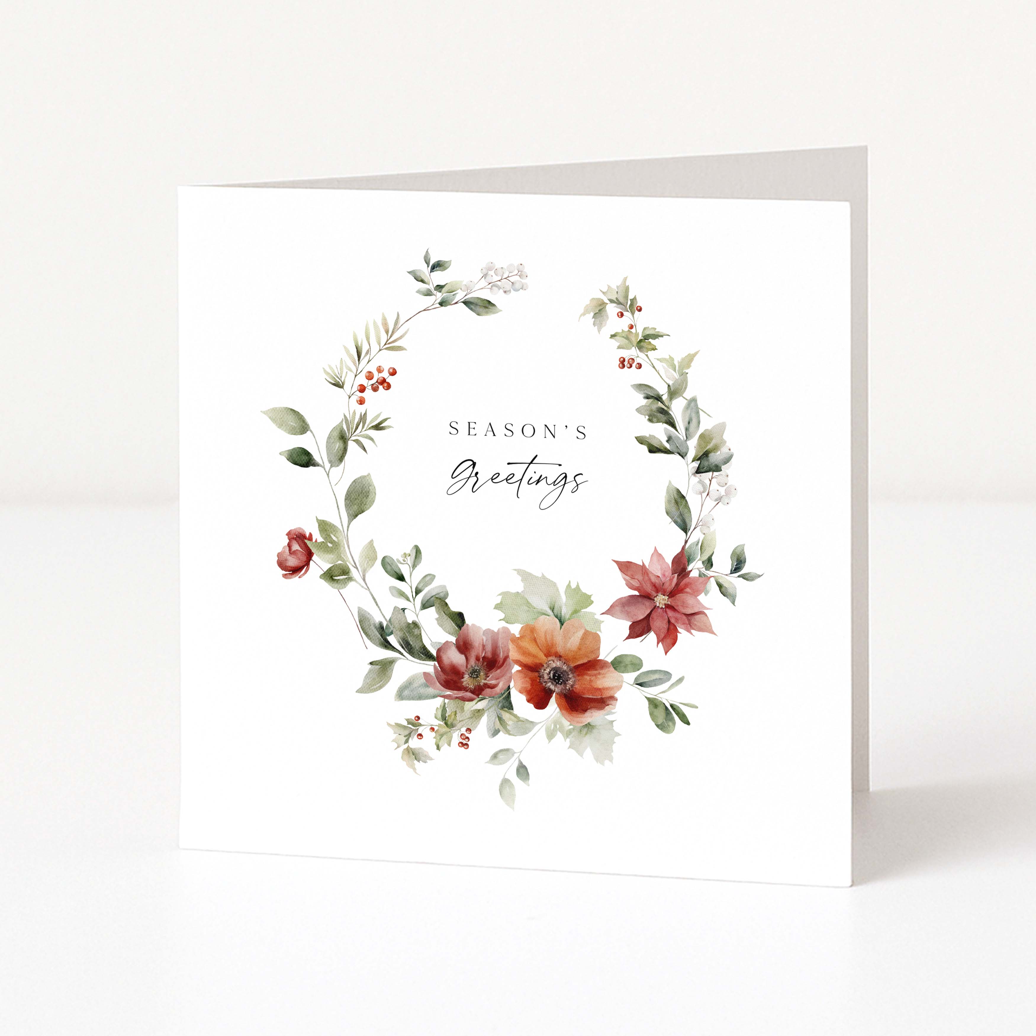 A white greeting card standing upright, featuring a watercolour floral wreath with green leaves, red and orange flowers, and the words 'Season's Greetings' in the centre.