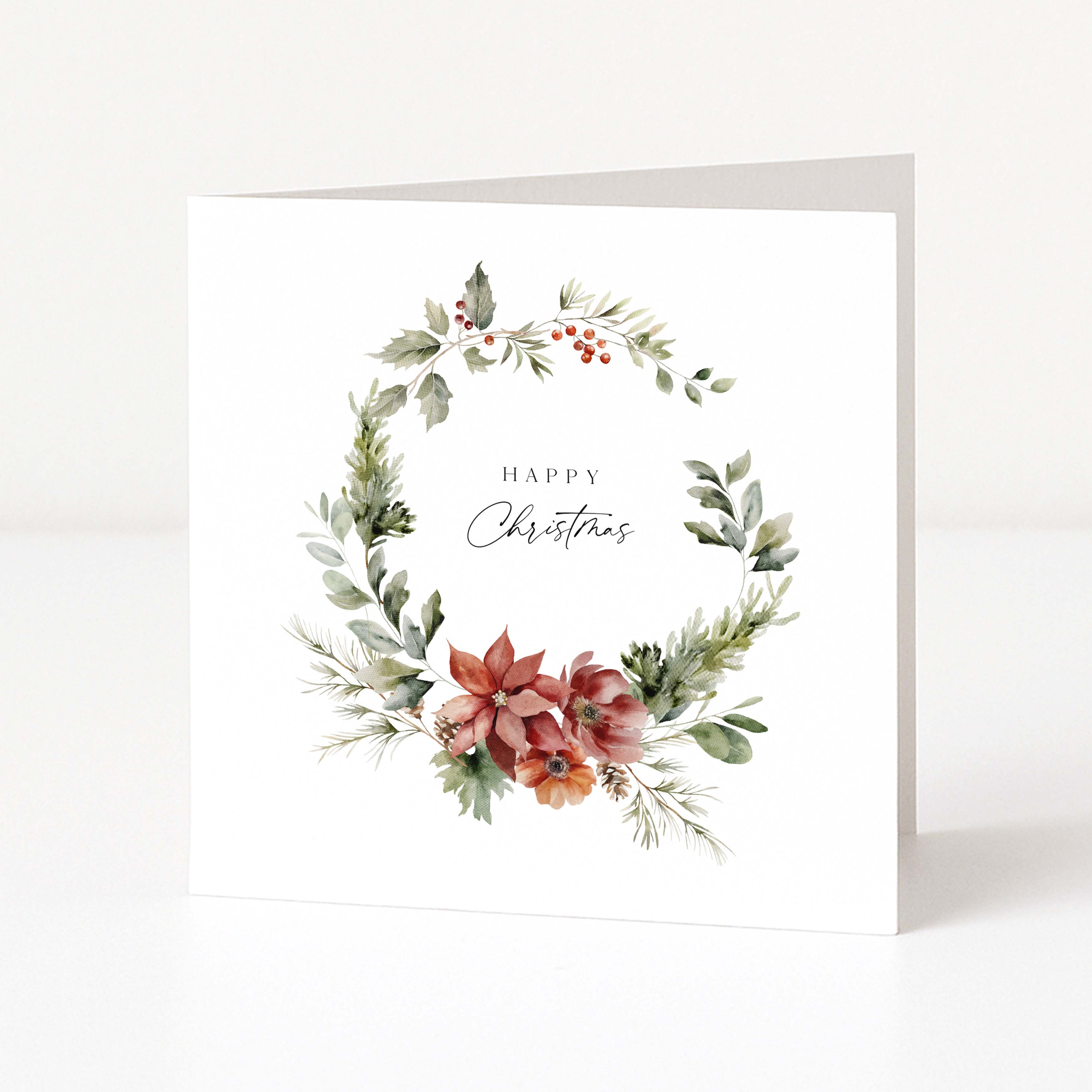 A square white Christmas greeting card standing upright, featuring a watercolour floral wreath with green leaves, red berries, and poinsettia-style flowers, with the words 'Happy Christmas' printed in the centre.