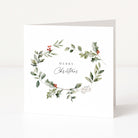 A white square greeting card standing upright on a light grey surface, featuring a watercolour wreath of green leaves and holly with small red berries and the words “MERRY Christmas” in the centre.