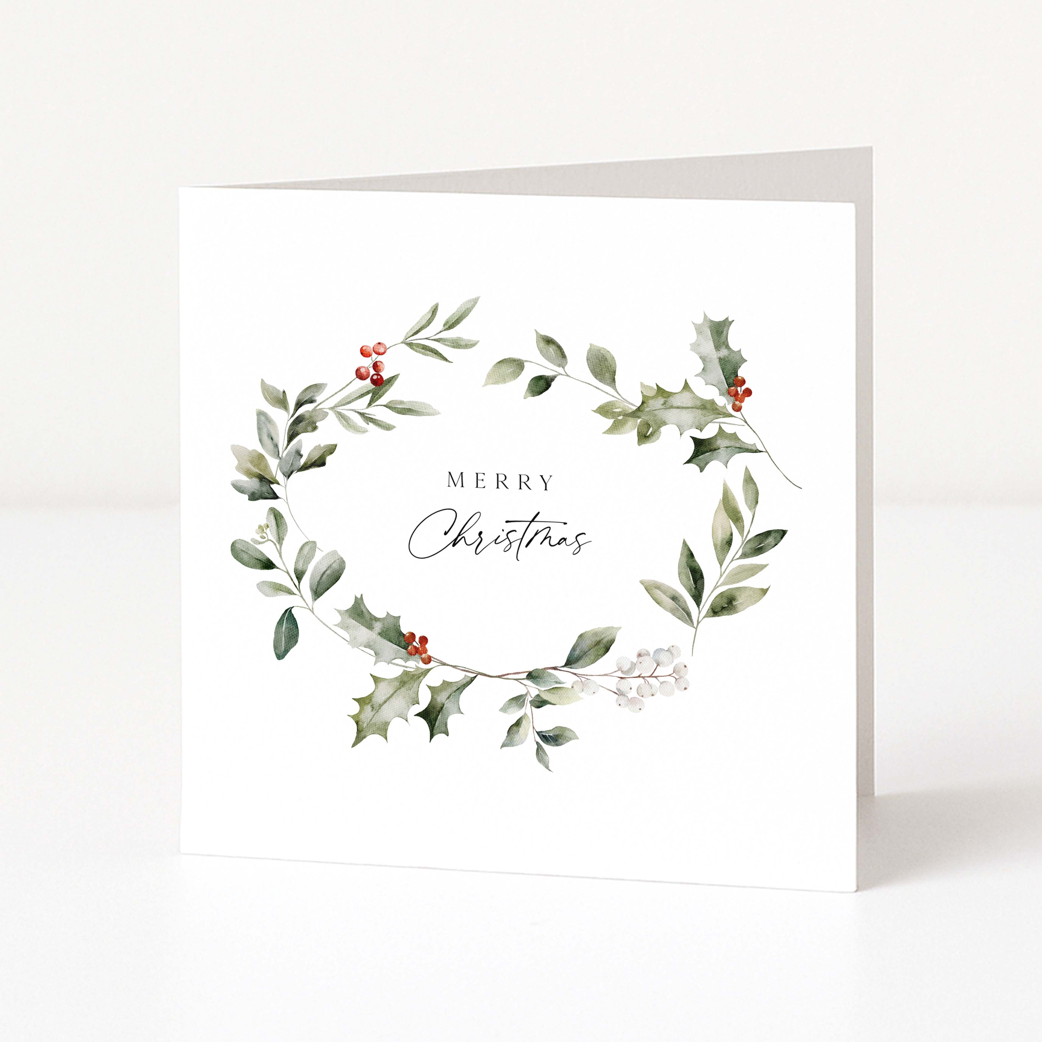 A white square greeting card standing upright on a light grey surface, featuring a watercolour wreath of green leaves and holly with small red berries and the words “MERRY Christmas” in the centre.