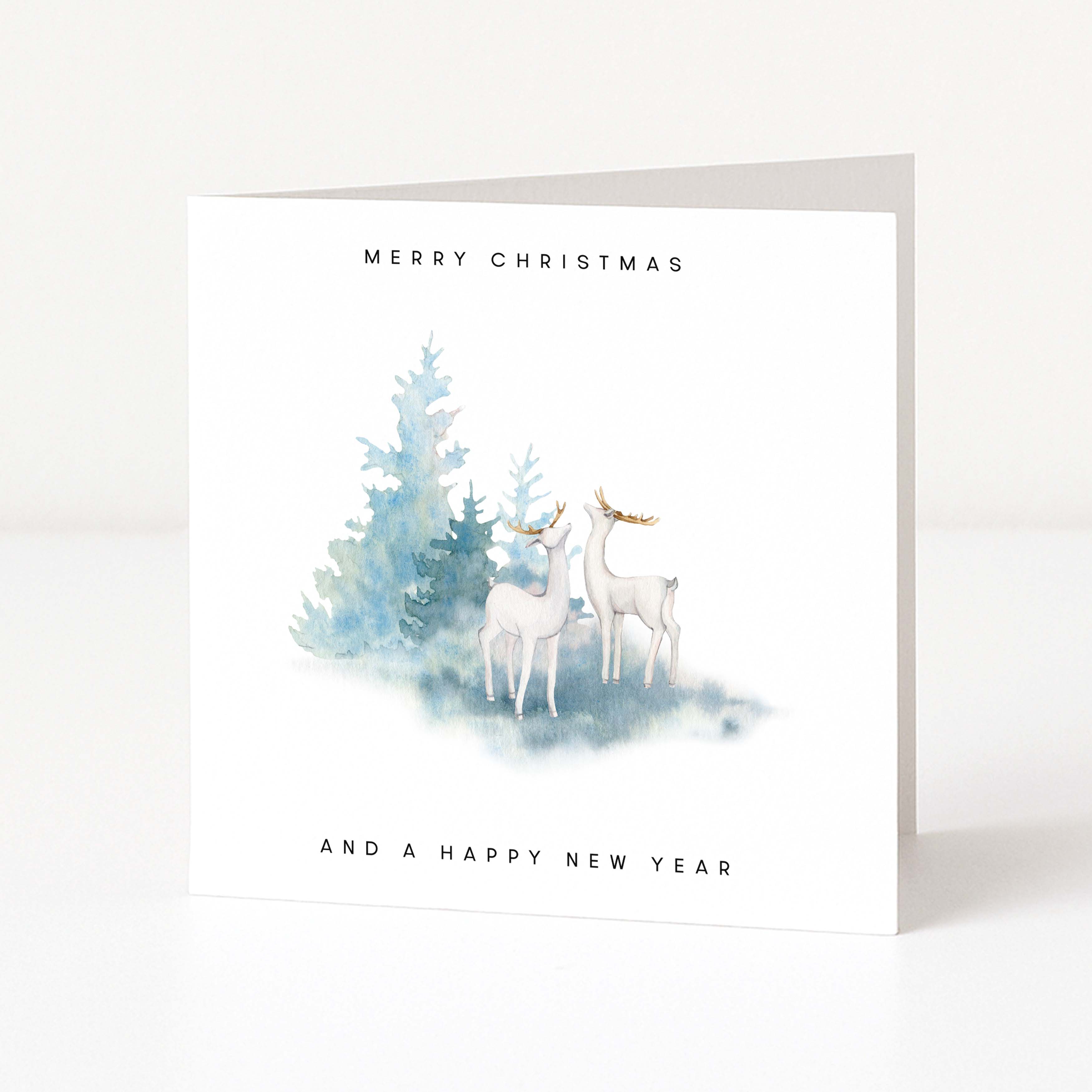 A white square greeting card standing upright on a light surface, printed with the text 'MERRY CHRISTMAS' at the top and 'AND A HAPPY NEW YEAR' at the bottom. The centre shows a soft watercolour illustration of two white deer beside blue-green evergreen trees.