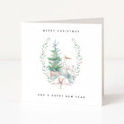 A white greeting card standing upright on a light surface, printed with the words 'MERRY CHRISTMAS AND A HAPPY NEW YEAR' around a watercolor illustration of a small tree, a deer, and pale botanical accents.